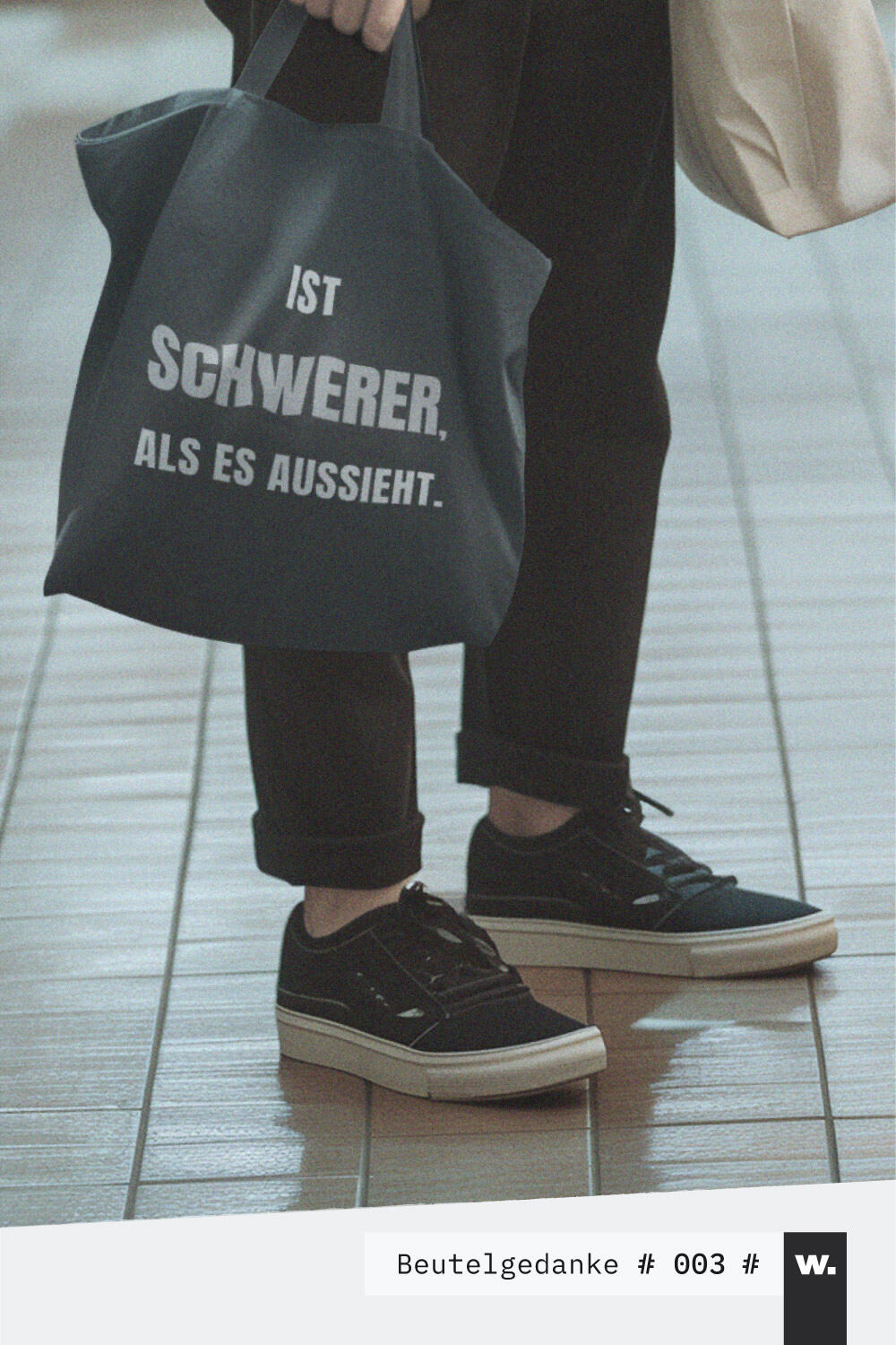 Jemand trägt einen schwarzen Stoffbeutel mit dem plakativen Aufdruck „Ist schwerer als es aussieht.“ in ruhiger Straßenszene.