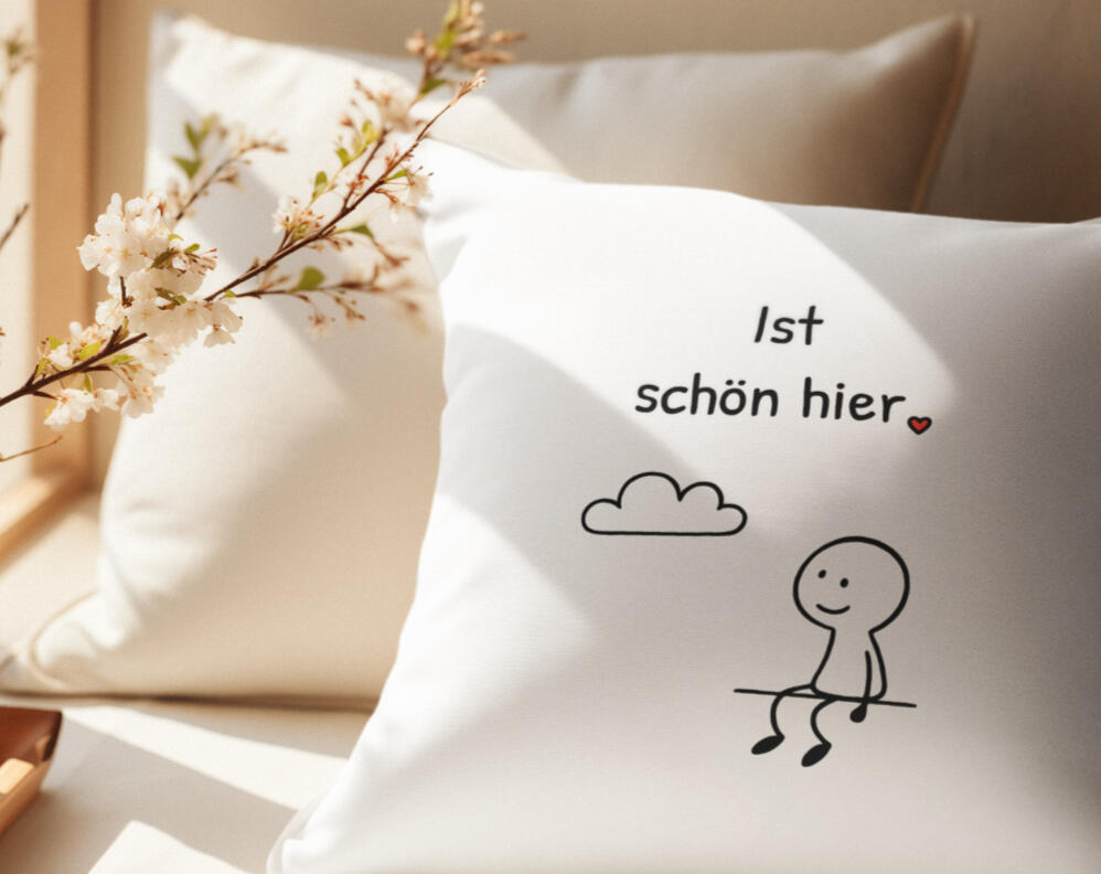 Helles Sofa-Kissen, bedruckt mit freundlicher Strich-Figur und einer Wolke mit dem Satz „Ist schön hier“ und kleinem roten Herz in ruhiger, gemütlicher Wohnszene, als kleine Geschenkidee mit Herz und schlichter Gestaltung.